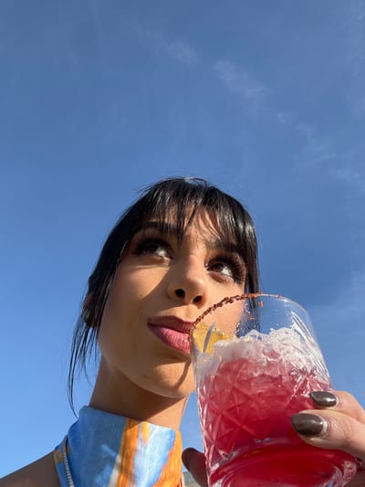An extreme low-angle shot captures a person holding a red drink against a bright blue sky. The subject, a person with dark hair and bangs, is viewed from below and slightly to their right. They have striking eye makeup, featuring long eyelashes and defined brows, and their lips are painted in a muted pinkish-brown shade. Their dark fingernails are visible as they hold the glass.

The drink is in a clear, faceted glass, filled with a vibrant red beverage, ice, and a lemon slice. The rim of the glass is coated with a brownish, granular substance, likely a sugar or salt rim. A portion of a colorful top, patterned with blues, oranges, and whites, is visible on the person's right shoulder. The person's gaze is directed upwards and slightly to the left, engaging with the camera.

The background is a vivid, cloudless blue sky, suggesting bright, clear weather. The strong overhead sun creates subtle shadows on the person's face and emphasizes the details of the drink. The image is a stylized portrait set outdoors on a sunny day in El Tomate, Mexico. There is no visible text in the image.