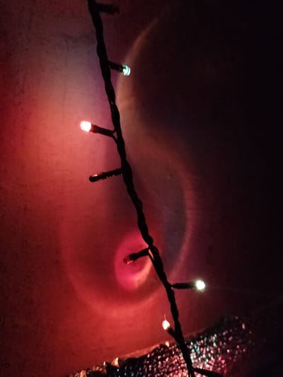 The image shows a string of festive lights draped vertically against a textured, reddish-pink background that appears to be a wall or fabric. The lights themselves are small and cylindrical, with some emitting a soft, warm white glow, while others are tinged with red or green hues. A circular, halo-like effect is visible in the upper right portion of the background, possibly caused by a light source or lens flare. The overall impression is one of low-light, atmospheric illumination, suggestive of a decorative setting, possibly a celebration or holiday. There are no people or specific landmarks visible to indicate a particular location within Kudashivka, Ukraine.