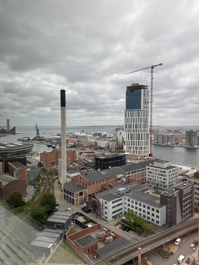 This is a high-angle, wide-angle shot of the Aarhus harbor area in Denmark, captured on a cloudy day. The scene is dominated by industrial and construction elements, with a clear view of the waterfront.
In the foreground and mid-ground, a variety of buildings are visible. To the left, a large, cylindrical industrial chimney with a black cap stands prominently. Several brick buildings, some with multiple stories and numerous windows, are clustered together. A modern, multi-story building with a dark facade and vertical louvers is also present. Adjacent to these, a curved, modern structure with a glass roof can be seen. A network of train tracks runs through this part of the scene, with a few parked cars and some greenery.
Dominating the mid-ground and extending towards the background is a very tall, modern skyscraper, partially under construction. It features a distinctive white and dark blue patterned facade. A large red construction crane is attached to the side of this building, with its arm extended over the structure.
Further back, the harbor is visible with several boats and ships. A large cruise ship is docked to the right, and industrial facilities like silos and storage tanks are scattered along the waterfront. In the distance, across the water, low-lying land and a hazy skyline can be seen. The sky is heavily overcast with thick, gray clouds. The overall impression is one of a working port city with ongoing development. There are no identifiable individuals clearly depicted in the image.