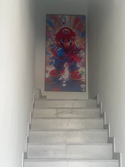 The image shows a narrow staircase leading upwards, flanked by plain white walls. Mounted on the wall above the staircase is a tall, narrow framed artwork featuring a stylized depiction of the character Mario from the Super Mario video game franchise. Mario is rendered in a vibrant, abstract style with splashes of red, blue, yellow, and pink paint. The artwork is positioned centrally on the wall. The staircase itself appears to be made of concrete or stone, with each step having a light grey tiled surface. The overall environment suggests an interior residential or commercial space. The specific location of Jinyun County, China, is not evident from the visual information in the image.