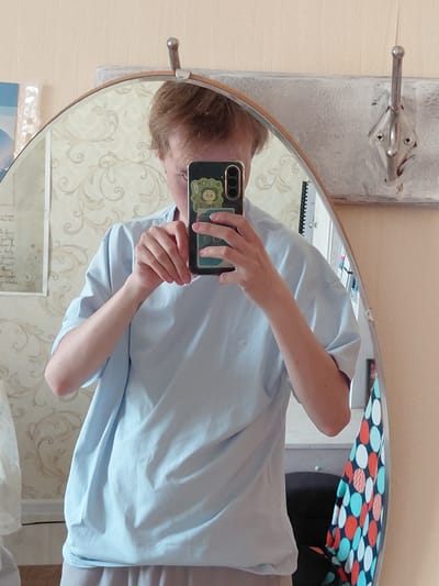 An individual is captured in a mirror selfie, with the image rotated 90 degrees counter-clockwise. The person, with short light brown hair, is wearing a light blue short-sleeved t-shirt and light-colored pants or shorts. They are holding a dark-colored smartphone with a light green/yellow cartoon-themed case, featuring an animal character, to take the photo. Their face is partially obscured by the phone.
The mirror, which is oval or rounded rectangular with a light wooden frame, reflects an indoor setting, likely in Nadym, Russia. The background features a wall covered in light-colored wallpaper with a subtle, swirling floral pattern. To the upper left of the mirror, a silver, two-pronged clothes hook is mounted on a textured, light grey wall panel. In the reflection, within the upper right curve of the mirror, a vibrant fabric with a pattern of red, white, and teal polka dots or circles is visible. Below the mirror, some white papers or plastic sheets with unreadable text or graphics are present. The overall scene depicts a casual indoor moment.