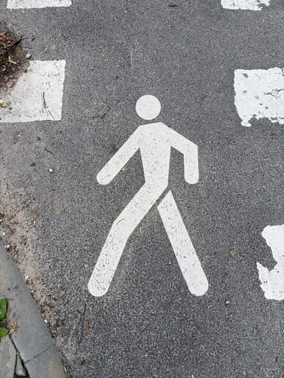 The image shows a white pedestrian crossing symbol painted on asphalt. The symbol depicts a stylized human figure with its arms outstretched and one leg forward, indicating movement. The symbol is worn and chipped in places, suggesting it is well-used. The asphalt surface is dark grey and speckled with small stones, characteristic of road surfaces. There are also several white rectangular markings, likely part of the pedestrian crossing, visible in the frame. Some debris, including leaves and small twigs, is scattered on the asphalt and at the edge of the curb. The curb itself is made of concrete. The image is taken from a high angle, looking directly down at the ground. The lighting appears to be natural daylight, casting no strong shadows, suggesting an overcast or diffused light condition. No specific time of day, weather, or emotional cues are evident. The image is a close-up, focusing entirely on the ground markings and the road surface. There is no visible text in the image.