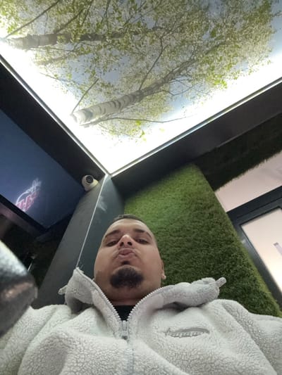 This image depicts a male individual taking a selfie indoors in Stornara, Italy. The subject is an adult male with short dark hair, a dark goatee, and an earring visible in his left ear. He is wearing a light-colored, possibly white or light grey, zippered fleece jacket over a dark undershirt, and is looking directly at the camera with his lips pursed. The immediate environment includes a prominent rectangular ceiling panel that functions as an illuminated display, showing an image of birch trees with light green leaves against a bright sky. To the subject's right, a wall is covered with artificial green turf. To his left, a dark vertical support structure or wall section features a small white dome-shaped security camera mounted near the top. Below this camera, a portion of a digital display with red graphics is visible. A dark-framed opening, possibly a window or doorway, is partially visible in the background to the right.