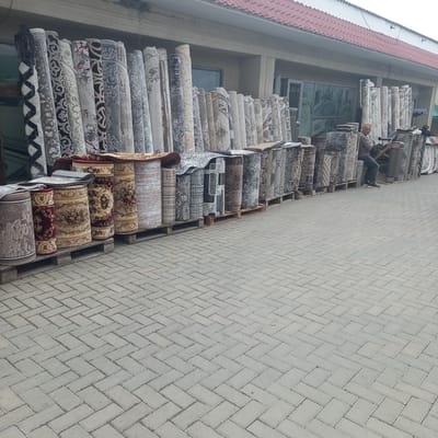 The image shows an outdoor carpet shop in Briceni, Moldova. The shop displays a large variety of rolled-up carpets, stacked on wooden pallets and arranged along the facade of a building. The carpets exhibit diverse patterns and colors, ranging from intricate traditional designs to more modern geometric prints. In the mid-ground, a man, dressed in a gray long-sleeved shirt and dark pants, is seated on a low structure, appearing to be a vendor or employee of the shop. He is looking towards the camera. The ground in front of the shop is paved with grey interlocking bricks arranged in a herringbone pattern. The building itself has a light-colored facade, with a red tiled roof visible at the top of the frame. Large windows and doors are visible on the building, some reflecting the surrounding environment. The scene suggests a commercial area on a possibly overcast day, with the focus on the extensive display of carpets.