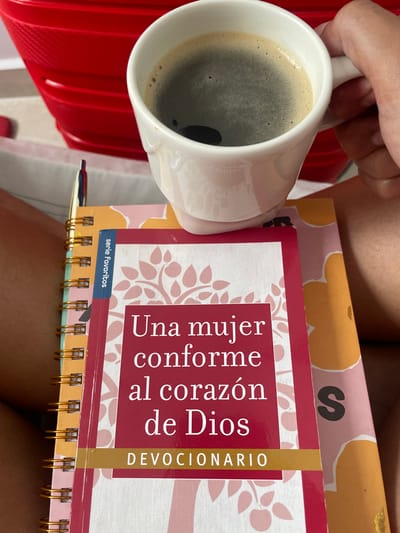 The image shows a white mug filled with a dark beverage, likely coffee, being held in a person's hand. The mug is positioned next to a book titled "Una mujer conforme al corazón de Dios" (A woman after God's own heart), part of the "serie favoritos". The book is a devotional and has a spiral binding. Beneath the book there's another with flowered pattern. A pen lays besides these. The setting is inside, possibly a home, but there are no contextual clues to place it specifically in Rionegro, Colombia. No further details about people, interactions, or environmental specifics are discernible from the image.