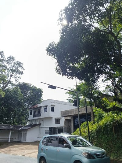 A light blue car is driving on a road in front of a two-story white building. A smaller white structure is connected to the building. A traffic light stands near the building, with foliage visible in the background. Trees partially obstruct the sky, with the sun visible through the leaves. Dense greenery covers the hillside to the right of the building.