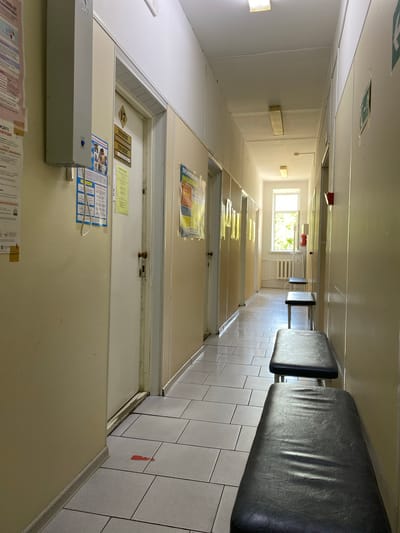 The image depicts a long, institutional hallway, possibly within a clinic or hospital in Zapadny, Russia. The hallway is brightly lit, suggesting daytime. On the right side of the frame, two dark, cushioned benches are mounted on the wall, facing into the hallway. The floor is tiled with light-colored squares. Along the left wall, there are several closed doors, some with signage. One door, labeled with the number "17", has two signs next to it, one of which reads "ТЕРАПЕВТ" (Therapist). Various posters and informational flyers are attached to the walls, particularly near the doors. One poster above the door labeled "17" appears to be a colorful informational chart. Further down the hallway, a window provides a view of green foliage, indicating trees or bushes outside, bathed in sunlight. Near the window, a radiator is visible. The overall impression is one of a clean, functional, and perhaps somewhat dated public building. There are no people visible in the image. The lighting suggests it is daytime, and the presence of foliage outside implies a temperate climate.