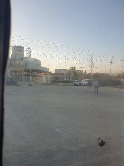 A sideways landscape shows a construction site with large cylindrical tanks and scaffolding under a pale sky with power lines. A road with a dark spot dominates the foreground. A person walks near parked cars in the middle ground, and low vegetation lines the back.