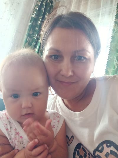 A selfie shows a woman and a baby. The woman, with brown hair and dark eyes, is smiling. She is wearing a white t-shirt. The baby, with blonde hair and blue eyes, is looking at the camera. The baby is wearing a white onesie with pink stars. They are indoors, with curtains in the background. The location is identified as Идрисово, Russia.