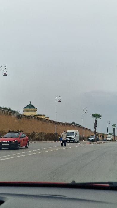 The image displays a street scene in Mechouar Fes Jdid, Morocco, under an overcast sky. A prominent light-brown fortified wall extends across the background from left to right. Behind this wall, a building featuring a light yellow facade and a distinctive green-tiled pyramidal roof is visible. The foreground and midground are dominated by a multi-lane asphalt road.
One individual, a male pedestrian dressed in a light-colored jacket and dark trousers, is situated centrally in the road, appearing to cross or stand. A red compact car with a roof rack is positioned in the left lane, facing away from the viewer. Further down the road, in the right lane, a white Mercedes-Benz van is stationary. Several other dark-colored vehicles are visible parked or stopped along the right side of the road, beyond the van. Black, ornate street lamps with double curved arms line the road, and several tall palm trees are visible along the right side of the road. The photograph appears to be taken from inside a vehicle, with a dark-red dashboard element visible at the bottom of the frame.
