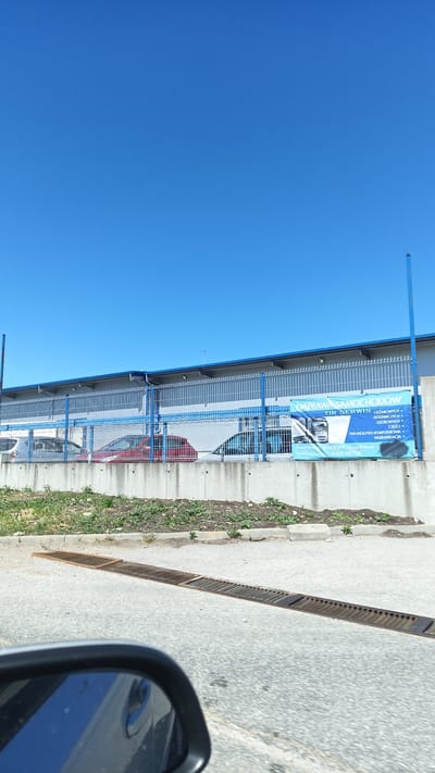 The image shows an automotive repair shop. A sign in Polish advertises "NAPRAWA SAMOCHODOW TIR SERWIS," which translates to "Truck Repair Service". The sign also lists services, including repairs for cars and vans, parts, computer diagnostics, and vulcanization. Contact information on the sign reads "POLEK MOTOREKULA, Osipy Kolonia 5, 664 189 920".
A light blue fence and gate with blue support posts secure the repair shop. Behind the fence, several cars are visible. A concrete retaining wall holds a small strip of soil and sparse vegetation. A metal drainage grate runs across the roadway in front of the shop. A car side mirror is visible in the foreground. The sky is clear and blue. The photo appears to be taken from a passing vehicle. The city referenced in the prompt, Plodovitovo, Bulgaria, is not present in the sign text. "Osipy Kolonia 5" appears on the sign, which is likely the address of the auto shop.