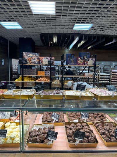 This is a portrait-oriented photograph taken inside a confectionery shop, likely in Russia. The scene is dominated by displays of sweets and baked goods. In the foreground, glass display cases reveal various chocolates, meringues, and possibly Turkish delight in trays. Some items are labeled with prices in Russian rubles. Behind these cases, shelves are stocked with boxed chocolates and other packaged sweets, also bearing Russian text. A person wearing a white apron and cap is visible in the middle ground, tending to the displays. The shop's interior features a modern grid-like ceiling with integrated lighting, and wooden shelving units. The overall atmosphere is that of a well-stocked, artisanal food shop. Visible text includes product names like "ЛУКУМ ШОКОЛАДНЫЙ" (Chocolate Loukoum), "ЗЕФИР" (Zephyr), and "МАРМЕЛАД" (Marmalade), along with prices such as "850" and "700". The lighting suggests it could be daytime or an indoor evening setting.