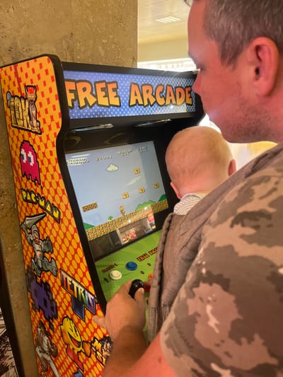 An adult male is operating a "FREE ARCADE" machine in an indoor setting within Vilobí d'Onyar, Spain. He is positioned facing the arcade machine, holding a young child in front of him, who also faces the screen. The man, wearing a camouflage-patterned short-sleeved shirt, has his right hand on the machine's black joystick and his left hand resting on the control panel near the white, blue, and orange buttons. The arcade screen displays gameplay from Super Mario Bros., featuring Mario in a landscape with question mark blocks and a cloud. The arcade cabinet's side art features characters and logos from classic games like Pac-Man and Tetris. A light-colored, possibly marble, pillar is visible to the left of the machine. The background beyond the subjects appears brightly lit.
