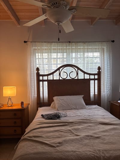 This image depicts the interior of a bedroom located in Olde Yard Village, British Virgin Islands. The scene presents a queen or full-sized bed with a distinctive wooden headboard, possibly constructed from rattan or bamboo, featuring decorative dark-colored scrolled elements at its apex. The bed is made with a white fitted sheet, a light-colored (beige or tan) blanket partially covering the mattress, and a single white pillow resting against the headboard. On the blanket, near the foot of the bed, a pair of dark-patterned shorts or pajamas and another light-colored garment are folded.
Behind the bed, a large window spans the wall, covered by translucent white sheer curtains, through which diffused daylight and indistinct outlines of exterior vegetation are visible. To the left of the bed, a three-drawer wooden nightstand, light brown in color, supports an illuminated table lamp with a square, off-white shade and a silver-toned base. Adjacent to the lamp, a red and white beverage can rests on the nightstand. A partial view of a similar wooden nightstand is visible on the far right of the frame, with a dark-colored object, possibly a mobile device, on its surface.
The room's ceiling features exposed light-colored wooden beams and planks. A white, four-bladed ceiling fan is centrally mounted, suspended from the wooden ceiling. The walls are light-colored, possibly beige or off-white. No individuals are present in the scene.