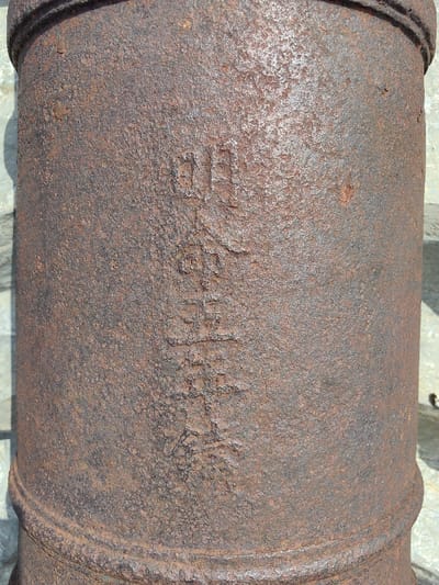 A close-up, vertical shot captures a heavily rusted cast iron object, presumed to be a section of an antique cannon barrel, located outdoors in Phú Xuân Ward, Vietnam. The cylindrical object exhibits a deeply pitted, rough, and oxidized surface, predominantly reddish-brown with darker grey patches, indicating extensive weathering. Raised characters, consistent with traditional Asian script, are vertically cast into the surface, partially obscured by corrosion but discernible. A circumferential raised band is present near the bottom of the visible section. Direct sunlight illuminates the object's right side, creating a distinct shadow line along its left. The blurred background consists of a light-colored, possibly paved or stone surface. No people, actions, or specific interactions are depicted.