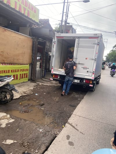The image depicts a man in a black shirt and blue jeans standing next to a white truck. The truck is parked on the street in Baleendah, Indonesia, at the coordinates -7.021, 107.657. The truck has its rear doors open and the man is facing the camera. There is a sign in the background that reads "Rumah Makan Padang Na Jaya".  There is a motorcycle in the foreground, and a puddle of water in the street in front of the truck. The street appears to be paved, but is in disrepair. The buildings in the background are a dull color. The sky is overcast and there are power lines overhead. The scene appears to be located in an urban area, possibly near a commercial district.