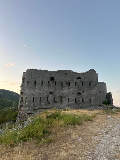 An ancient, stone fortress, likely abandoned and in a state of disrepair, stands on a grassy hillside. The fortress is constructed from large, grey stones and features numerous rectangular openings, suggestive of windows or gun emplacements. The structure has a rounded, imposing facade, with parts of the upper walls crumbled and overgrown with vegetation.
The fortress is situated in a rugged, natural environment. In the background, a lush green hillside slopes upwards, covered in trees and foliage. The sky above is a clear, pale blue, with a few faint clouds present near the horizon. The foreground consists of dry, yellow grass and rocky terrain, with a rough path made of gravel and stones leading towards the fortress.
This fortress is located in the Brajici area of Montenegro. The scene appears to be captured during the golden hour, with the soft light of dusk casting a warm glow. There are no people visible in the image, and the focus is entirely on the historic structure and its surroundings.