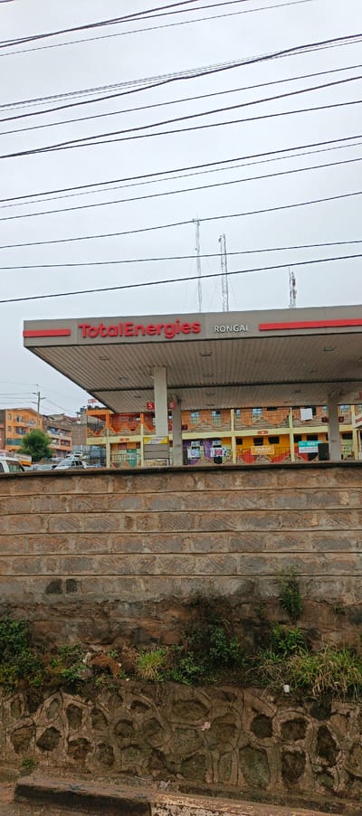 A Total Energies gas station in Rongai, is seen behind a layered brick wall. The sky is overcast with visible power lines above. The gas station is numbered 5 and 6. Beyond the station are colorful buildings. Cars are parked behind the wall. Vegetation grows at the base of the brick wall. There are three cell towers behind the gas station.