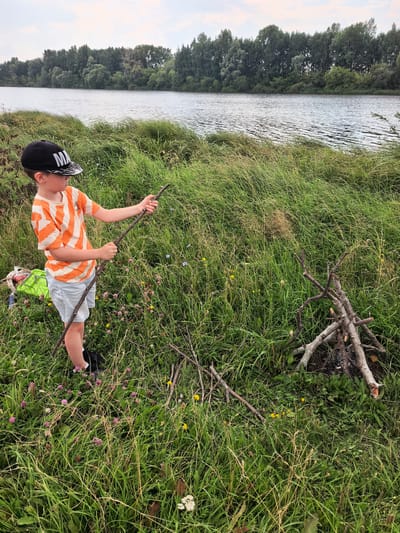 A young boy wearing a black baseball cap and an orange and white striped t-shirt stands in tall green grass next to a body of water. He is holding a long stick in his hands. In the background, a line of trees borders the water, and a cloudy sky is visible above. A pile of firewood is in the grass to the right of the boy. The scene appears to be outdoors in a natural setting.