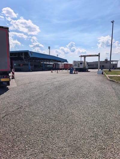 The image depicts a border crossing or inspection point, likely for freight trucks, under a bright, partially cloudy sky. The foreground is dominated by a grey, gravel-covered road. In the middle ground, a large blue-roofed structure, possibly a customs building or inspection shed, is visible, with several trucks parked or moving under it. To the right of the blue structure, a large, white gantry-like scanning device is positioned over the road, with a red "X" illuminated on its display. Various traffic cones and informational signs are present in the scene, indicating a controlled area. A tall pole with lights stands on the far right. The location is identified as Sid Municipality, Serbia.