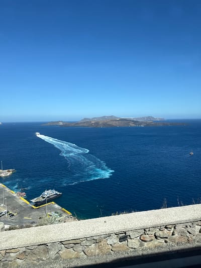 An eye-level outdoor shot captures a vast expanse of deep blue Aegean Sea under a clear, bright blue sky. In the mid-ground, a white speedboat is moving away from the viewer, leaving a prominent foamy white wake that stretches across the water. In the foreground, a gray stone wall with a rough, textured finish borders the bottom of the frame, with a smaller, lighter-colored wall built atop it. To the left, a pier juts out into the sea, with several boats docked. Among them, a larger blue and white ferry is moored. A few people are visible on the pier, and several cars are parked. In the distance, a chain of islands with rugged, mountainous terrain stretches across the horizon, with faint hints of white structures on the slopes. The scene appears to be in the port area of Thira, Santorini, Greece.