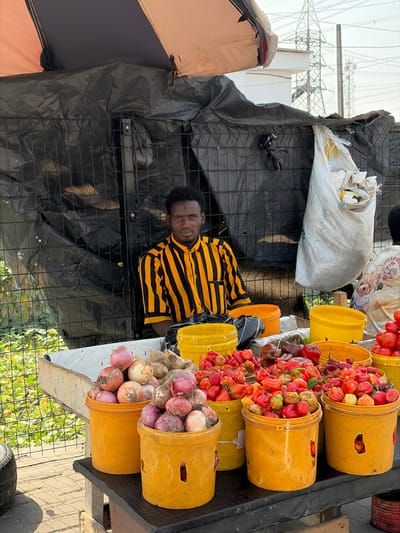 An adult male with dark skin and short black hair, wearing a yellow and black vertically striped short-sleeved shirt, is positioned behind a market stall in Jakande, Nigeria. He looks directly forward. The stall's backdrop consists of a dark, possibly black, tarpaulin or plastic sheeting secured to a metal grid fence. An overhead light-colored fabric awning provides shade. The counter in front of the individual displays an assortment of fresh produce arranged in multiple yellow plastic buckets. Prominently featured are buckets filled with red and white onions, and numerous buckets overflowing with red, orange, and green peppers, including many Scotch bonnet peppers. Additional buckets contain red tomatoes. A large white woven sack hangs from the background structure on the right. In the far background, large electricity transmission towers and a light-colored building are visible beneath a hazy sky. Green vegetation is present on the left side of the frame.