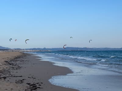 A wide, sandy beach runs along the left side of the image, meeting a vibrant blue ocean on the right. The sand is textured with dark patches, possibly seaweed or debris. A line of low buildings and mountains forms the background on the distant shore, suggesting an urban or coastal settlement. The sky is a clear, pale blue.
Several kite surfers are visible in the sky and on the water. One kite surfer with a red and black kite is in the mid-right section of the image, actively riding waves. Other kites, in colors of green, red, and blue, are scattered across the sky. A few indistinct figures can be seen on the beach in the distance, and a lighthouse is visible on the horizon. The scene depicts a typical day for kite surfing in Tarifa, Spain, a renowned location for this water sport.