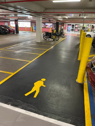 The image displays an indoor parking garage, likely part of a shopping mall or large commercial complex, evidenced by visible storefronts and display screens in the background. The viewpoint is slightly elevated, looking down an aisle of the parking facility.

The floor is dark grey or black, marked with bright yellow lines. On the left side, these lines delineate numerous parking bays specifically for motorbikes or scooters, with several black motorbikes parked. Each bay is numbered, such as 'M 169', 'M 170', and 'M 171'. A wide pedestrian pathway, also outlined in yellow, occupies the right side of the frame, featuring a large, yellow stencil of a walking person, clearly indicating a pedestrian-only area.

Several cars are partially visible. A maroon car and a white car are in the upper right, positioned behind yellow cylindrical protective bollards. A black car is seen towards the bottom left. The infrastructure includes numerous overhead fluorescent lights, red pipes, and conduits running along the ceiling. White concrete pillars support the structure, some of which feature digital display screens or signs with the text "EVENT." A small red "STOP" sign is also attached to one of the pillars.

The scene suggests an active and well-organized commercial parking environment. The lighting is entirely artificial and bright, typical for an enclosed garage. No individuals are present in the frame, but the designated pathways and parking spots imply regular use by both vehicles and pedestrians. The location is contextualized as Indooroopilly, Australia.
