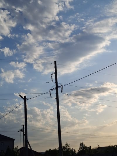 The image captures a sky filled with dramatic, wispy clouds against a backdrop of pale blue and a muted yellow sunset glow. Power lines crisscross the frame, silhouetted against the sky and supported by utility poles. One pole is prominently visible in the foreground, its structure a dark, angular shape. In the distance, along the right edge of the image, the dark shapes of trees and a building are visible, suggesting an urban or suburban setting. The overall mood is serene and somewhat contemplative, with the soft light of dusk casting a tranquil atmosphere. The image appears to be taken from the perspective of someone looking upwards, emphasizing the vastness of the sky and the intricate network of power lines. There is no visible text in the image.