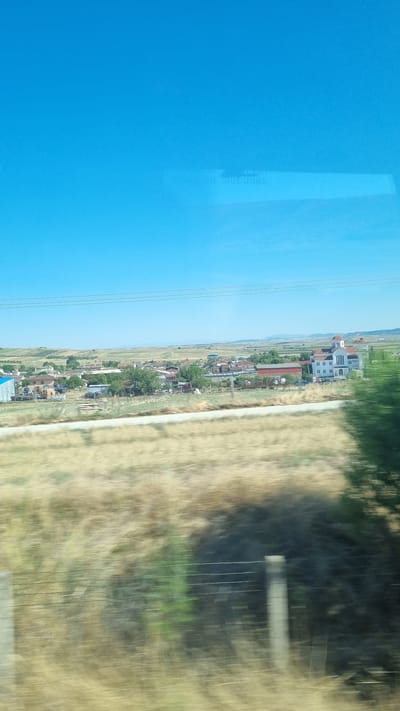 The image shows a village in the Municipal Unit of Kilada, Greece, on a sunny day. The sky is clear and blue. In the foreground, there is a field of dry grass and some blurry green vegetation, indicating movement, likely from a vehicle. A fence made of posts and wires runs horizontally across the middle ground. In the background, there is a cluster of buildings, including houses and a church with a white facade and a red-tiled roof. The village is surrounded by rolling hills and fields, with some trees scattered throughout. The overall scene depicts a rural landscape in Greece.