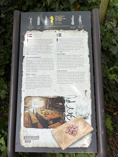 The image shows a sign providing historical information about the region. The sign is bilingual, written in both Dutch and English. It discusses the time of the Franks and Vikings, the role of the nobleman Sigiward, the rise of Christianity, and trade with the Friesians and Vikings. There is a photograph of the interior of an old building on the sign. The sign is on a pole surrounded by greenery, including some kind of ivy. A wooden cutting board with small sausages is laid on top of the sign. It appears to be located outdoors, near vegetation, specifically in Eindhoven, Netherlands.