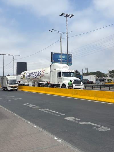 The image captures a busy roadside scene in San Juan de Miraflores, Peru, on a clear, cloudy day. Several trucks are visible on a paved road next to a yellow barrier.

In the foreground, a large white tanker truck belonging to "Llamagas" is prominently featured. The side of the tank reads "Llamagas," "Oriente," "GAS COMBUSTIBLE NO FUMAR" (Combustible Gas Do Not Smoke), "TACAR 14 200 GLNS," and the phone number "985 601 733." Directly in front of the tanker, a white tractor-trailer is parked, with "PELIGRO COMBUSTIBLE" (Danger Combustible), "Señor de Huanca," and "049 651 2" visible on its side. A person wearing a bright green safety vest is seen near the front wheel of this tractor-trailer, seemingly inspecting something or working. Further back, a smaller white cargo truck with the brand "FORLAND" on its cab is also present, along with other vehicles in the background.

The road features white painted markings, including what appears to be a large "S" or serpentine shape. Adjacent to the road is a sidewalk made of grey pavers. Overhead, power lines crisscross the sky, and multiple streetlights are mounted on poles. In the mid-ground, a large blue billboard displays text including "UNIDOS EN OBRA" (United in Work), "MIR IMINA," and "DE CALIDAD." The scene depicts typical urban or highway infrastructure, with vehicles potentially in transit or stopped for maintenance.