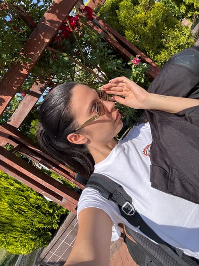 A young woman with dark hair tied in a ponytail, wearing sunglasses, a white t-shirt, and dark pants, is pictured outdoors. She is leaning back, with her hand near her face, and appears to be sitting on a bench. She is wearing a backpack. The background consists of a wooden pergola with red roses and green bushes. The scene suggests a sunny day, possibly in the city of Chavdar, Bulgaria.