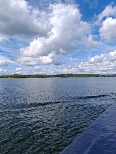 The image depicts a panoramic view of a large body of water under a partly cloudy sky, observed from a low vantage point on a vessel, likely in the Mörkö archipelago, Sweden. The upper half of the frame is dominated by a blue sky dotted with numerous white and grey cumulus clouds of varying sizes and densities. The lower half features expansive dark blue water, exhibiting widespread ripples and small waves across its surface, with more pronounced wave patterns visible in the immediate foreground, suggesting motion or current.

A distinct, forested landmass forms the distant horizon across the water, characterized by a dense line of green trees. In the mid-ground on the right side of the water, a single white sailboat with a visible mast and sails partially furled is positioned, appearing to be at anchor or slowly moving. The bottom right corner of the image shows a dark blue, textured surface, angled upwards from the bottom right, which is consistent with a railing or gunwale of a boat or ferry from which the photograph was taken. No individuals are visible.