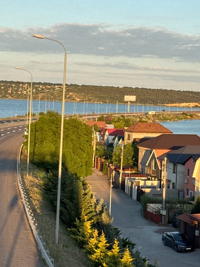 The image depicts a roadside view in Yelyzavetivka, Ukraine, with a highway on the left and a residential street on the right. A body of water, likely a river or bay, is visible in the background, with a distant treeline and a bridge or elevated road crossing the water. Several streetlights line the highway. The residential street features a row of houses with various architectural styles and roof colors, interspersed with trees and fences. A dark-colored car is parked on the right side of the street. The sky is clear with some scattered clouds, and the scene appears to be illuminated by the warm light of either sunrise or sunset.