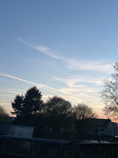 The image shows a sky with several contrails intersecting. The sky transitions from blue to a faint yellow-orange towards the horizon. Silhouetted trees and part of a building are visible to the right. A fence and what appears to be a greenhouse are also present. Based on the location information, this scene is likely in Würselen, Germany. There are no people visible.