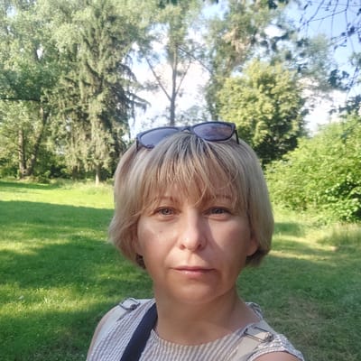 The media file is a portrait of a woman outdoors, taken at eye level. The woman has short, blonde hair and is wearing sunglasses pushed up onto her head. She is looking directly at the camera with a neutral expression. She is wearing a striped top. The background is a park with green grass and trees, suggesting a daytime setting on a sunny day. The shadows on the grass indicate that the sun is likely in the sky, casting shadows. The overall scene is peaceful and natural. There is no visible text in the image. The location is identified as Voronkiv, Ukraine.