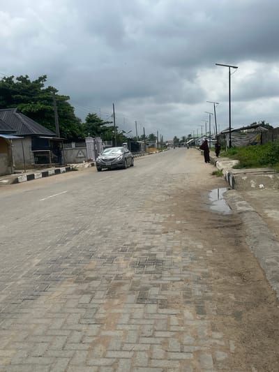 This image depicts a wide-angle view of a street in Mosafejaw, Nigeria, on an overcast day. The scene is dominated by a grey, cloud-filled sky above a residential and semi-commercial area.
The primary subject is a paved road, constructed with interlocking concrete pavers in a herringbone pattern, extending into the distance and appearing to curve slightly to the right. A faded white dashed line runs down the center of the road. On the left side of the road, a concrete curb painted with alternating black and white stripes delineates the road from a dirt shoulder. Beyond the shoulder, various buildings are visible, including a structure with a dark corrugated metal roof and grey walls, and another building partially obscured by a tree, featuring a white ornate gate and fence. A grey Hyundai Sonata sedan (license plate partially visible as "255 HDO") is parked or slowly moving on the left side of the road, facing away from the viewer. A lone figure in a dark blue top and pants is standing near the entrance of the dark-roofed building on the left. Electrical or utility poles with overhead wires run along the left side of the street.
On the right side of the road, the curb is less defined and transitions into a dirt and gravel shoulder, containing a notable puddle of water. Further along this side, two individuals are walking away from the viewer; one is wearing a dark, possibly brown, full-length garment and is closer to the foreground, while the second figure, dressed in similar dark clothing, is further down the road. A series of modern streetlights, characterized by tall, thin poles with flat, rectangular luminaires, are spaced along the right side of the road, extending into the distance. Behind the streetlights and people, a mix of simple structures, some with makeshift appearances, and dense green vegetation are visible. A large banner with text, including "200," is strung between poles on the right. In the far distance, other vehicles, possibly motorcycles or tricycles, are barely discernible. The ground on the shoulders is uneven and appears damp or muddy in places.