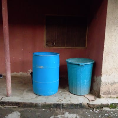The image shows two blue plastic containers, likely water barrels, positioned on a concrete ground. The larger barrel on the left is cylindrical and appears to be a standard water storage drum. To its right, a slightly smaller, wider barrel with a ribbed texture is also visible. Both containers are empty or nearly empty.
The scene is set outdoors, in what appears to be a covered area with a reddish-brown wall in the background. There's a narrow, dark opening or window above and between the barrels, covered with a wire mesh. A reddish-brown pillar is present on the left side of the image, partially obscuring a dark object on the ground that could be a pair of shoes.
The concrete ground is cracked and appears worn, with some patches of dirt and moss, suggesting an outdoor setting that might be prone to dampness. The wall itself shows signs of age, with peeling paint or plaster in some areas. The overall lighting suggests it could be daytime, possibly overcast, as there are no strong shadows.
The location context provided is Mangu Town, Nigeria. There is no visible text in the image. The focus of the image is on the two barrels and their immediate surroundings, hinting at a utilitarian space likely used for water storage or as part of household or community infrastructure. There are no people or animals visible. The mood is neutral, focusing on the objects and the environment.