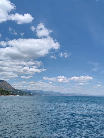 The image depicts a body of water with a coastline and mountains in the background, under a bright blue sky with scattered white clouds. The water is a deep blue and appears calm, with small ripples on its surface. The coastline is lined with trees and buildings, suggesting a populated area. In the distance, mountains form a hazy silhouette against the horizon. The scene is tranquil and appears to be a landscape view of a coastal city or town. The location is Utios, Ukraine.