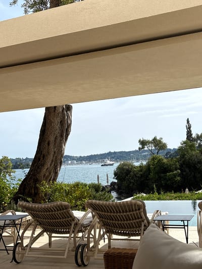 The scene is an outdoor terrace in Gouvia, Greece, overlooking the sea. The terrace features wicker lounge chairs with wheels and small tables. A thick tree trunk extends from the foreground towards the sea. The sea is visible in the background, with a coastline lined with buildings and vegetation. The sky is partly cloudy.
