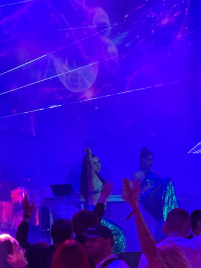 The image depicts an indoor event with a crowd of people, likely a concert or performance. The scene is dominated by vibrant blue and purple lighting, suggesting a club or stage setting. On a raised platform, two individuals, presumably performers, are visible. The performer on the left is dressed in a shimmering blue sequined outfit, with one arm raised. The performer on the right is also in a sequined outfit and appears to be interacting with the audience. In the foreground, several audience members can be seen, with their hands raised, indicating engagement with the event. A laptop and musical equipment are present on the stage, suggesting a musical performance. The background features abstract light projections and laser beams, contributing to the energetic atmosphere. The city of Akhalubani in Georgia is not explicitly indicated in the image, and thus cannot be inferred or confirmed.