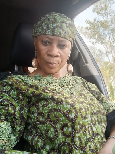 A woman is shown in a close-up shot while inside a car. She has brown eyes, dark hair, and appears to be wearing a green and brown patterned outfit with matching headwear. Large gold earrings are visible. The location is inferred to be Rigachikun, Nigeria, based on the prompt's request, but there are no distinct landmarks or features to confirm this. Outside the car window, trees and sky are visible. The woman is positioned in the driver's seat.