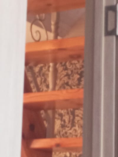 The image depicts an out-of-focus, partial view of an indoor wooden staircase, framed by a light-colored vertical element on the left and a darker, possibly metallic, vertical frame with a dark rectangular attachment on the right. Multiple light brown wooden stair treads are visible, ascending towards the top right of the frame. A dark, scrolled metal decorative element, likely part of a balustrade or railing, is discernible between two upper treads. Behind the staircase, a wall or surface with a dark, intricate, and textured pattern is visible. No individuals or specific actions are present. The image provides no discernible features or geographical markers to confirm the location as the city of Яромаска, Russia.