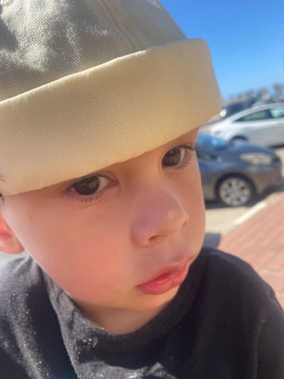 The image shows a close-up, low-angle shot of a baby's face. The baby is wearing a light-colored beanie that covers part of their forehead and eyes. The baby's skin is smooth and rosy, and their lips are parted, revealing a hint of teeth. A dark hoodie is visible on the right side of the frame, with some glittery details. The background is blurred, showing a street scene with a car and a blue sky. The lighting suggests it is daytime, likely sunny, given the bright illumination. The overall impression is one of innocence and curiosity. The text "Kiryat Yam, Israel" appears as a location context.