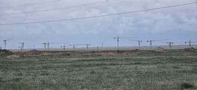 The image depicts a construction site with numerous tower cranes visible against a cloudy sky. The cranes are spaced out across a barren, grassy plain, suggesting an expansive project. In the mid-ground, a dirt embankment runs horizontally across the scene, with a small, dark-roofed building situated on top of it. The terrain appears undeveloped, with piles of earth and some fencing visible. Overhead, power lines stretch across the frame. The scene is devoid of people or vehicles. The location is identified as the city of Koshkol', Kyrgyzstan, indicating a construction project underway in a rural or semi-rural setting.