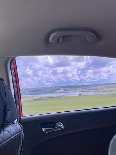 The image captures a view from the interior of a vehicle, looking out a window. Inside the car, the grey fabric of the headliner is visible at the top, with a plastic grab handle attached. Below this, a portion of a black quilted seat cover is seen on the left. The door panel, made of black plastic with a textured surface, is prominent in the lower right, featuring a silver door handle. Through the window, a landscape unfolds under a sky filled with white and grey clouds. The scene outside consists of rolling green hills, a body of water that appears to be a river or lake, and scattered trees. The overall impression is of a travel scene, possibly on a road trip, with the car interior framing the expansive natural scenery. The location in the city of Tolbazy, Russia, is not explicitly evident from the visual cues in the image alone.