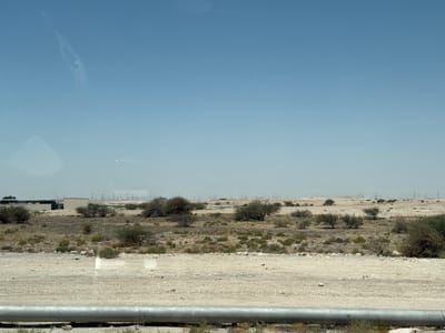 The image displays an expansive, arid desert landscape under a clear, pale blue sky, likely captured from inside a vehicle given the reflections and glare visible in the upper left portion of the frame. In the immediate foreground, a large, grey metal pipe runs horizontally, partially obscured by sparse, dry vegetation and gravelly soil.

The midground and background consist of vast stretches of rocky, dusty ground interspersed with scattered, low-lying desert bushes and small, resilient trees. Towards the distant horizon, an industrial area is evident, featuring several light-colored buildings and numerous power transmission towers or similar utility structures extending into the haze.

No specific activity or event is taking place; the scene is a static landscape view. Notable details include the bright daytime lighting, suggesting a clear and possibly hot day, and the characteristic desert flora. The presence of industrial infrastructure indicates human development within this natural environment. No people or visible text are present. The scene aligns with the geographic context of Umm Salal Ali, Qatar.