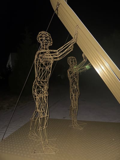 This media file features a wireframe sculpture of a person in a dynamic pose, appearing to swim or fly. The sculpture is metallic, likely made of brass or a similar alloy, and is illuminated by an unseen light source that casts subtle shadows. A reflection of the sculpture is visible on a polished surface below, giving the impression of a dual image.
The background is dark, suggesting it is nighttime or a dimly lit indoor space. Part of the scene includes a light-colored, curved overhead structure that casts a shadow on the sculpture and its reflection. The ground beneath the sculpture appears to be a textured, metallic surface, possibly a diamond plate or similar industrial flooring.
The sculpture itself is intricate, with each limb and the torso formed by a network of thin, connected wires creating a skeletal outline of a human figure. The figure's arms are raised, with one extended forward and the other bent, as if in motion. The legs are also extended, contributing to the sense of movement.
There are no visible text elements in the media file. The overall mood is one of artistic display, with the lighting and reflections enhancing the visual appeal of the sculpture. The context of Egorlykskaya, Russia, does not provide specific visual cues within the image itself, as the scene appears to be a general art installation.