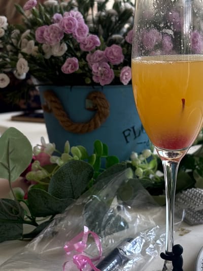 The image shows a close-up of a champagne flute filled with an orange liquid and a single red berry. The flute rests on a white tablecloth. In the background, a blue bucket with rope handles contains a bouquet of pink and white artificial flowers. To the left and right of the flute, there are more artificial green leaves and pink flowers. In the foreground, a clear plastic wrapper with pink ribbon and a black tube are visible. A small black figurine is clinging to the stem of the champagne flute. The scene appears to be indoors or in a sheltered outdoor setting, given the artificial flowers and tablecloth. The location is not discernible from the image itself, and no information about Santiago Yancuitlalpan, Mexico, can be inferred.